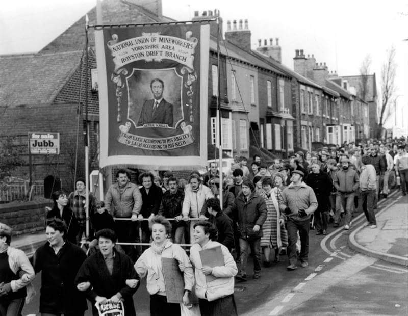 A black and white photograph of a march featuring members of the National Union of Mineworkers carrying a large banner with a portrait of Arthur Scargill, surrounded by a crowd of supporters along a street.