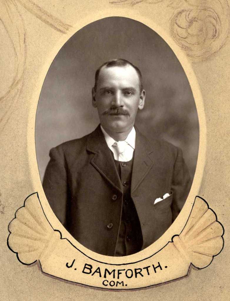 A black and white portrait photograph of a man with a mustache, dressed in a formal suit with a bow tie. The image is framed with decorative elements and the name 'J. Bamforth' is displayed at the bottom.