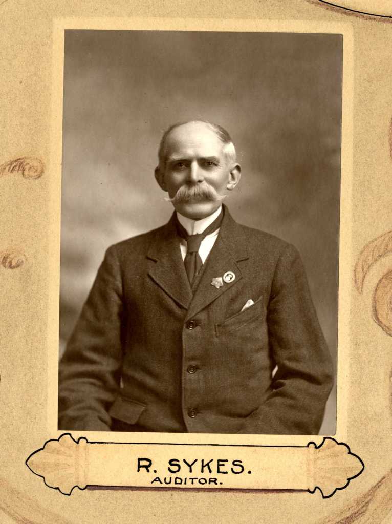 Black and white portrait of a man named R. Sykes, identified as an auditor, wearing a suit with a pocket square and a tie.
