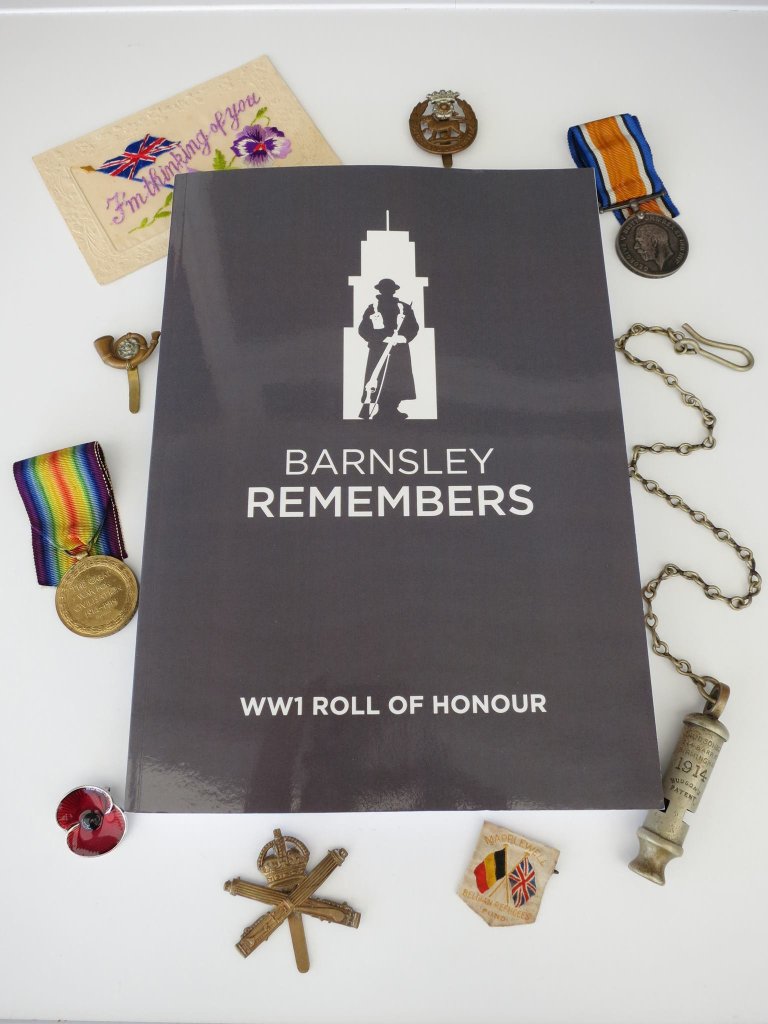 A flat lay photo featuring the 'Barnsley Remembers WW1 Roll of Honour' book surrounded by various military medals, badges, and a remembrance poppy.
