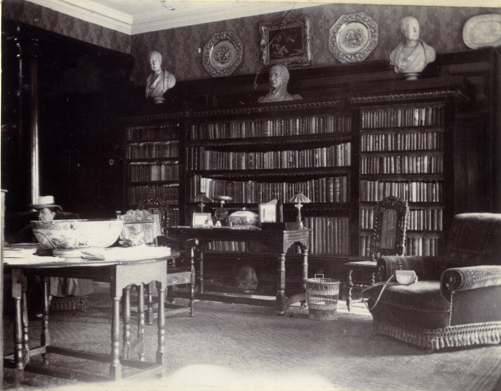 Interior view of a library room featuring bookshelves filled with books, a decorative bowl on a table, a lamp, and ornate furniture, capturing a glimpse of Edwardian domestic life.