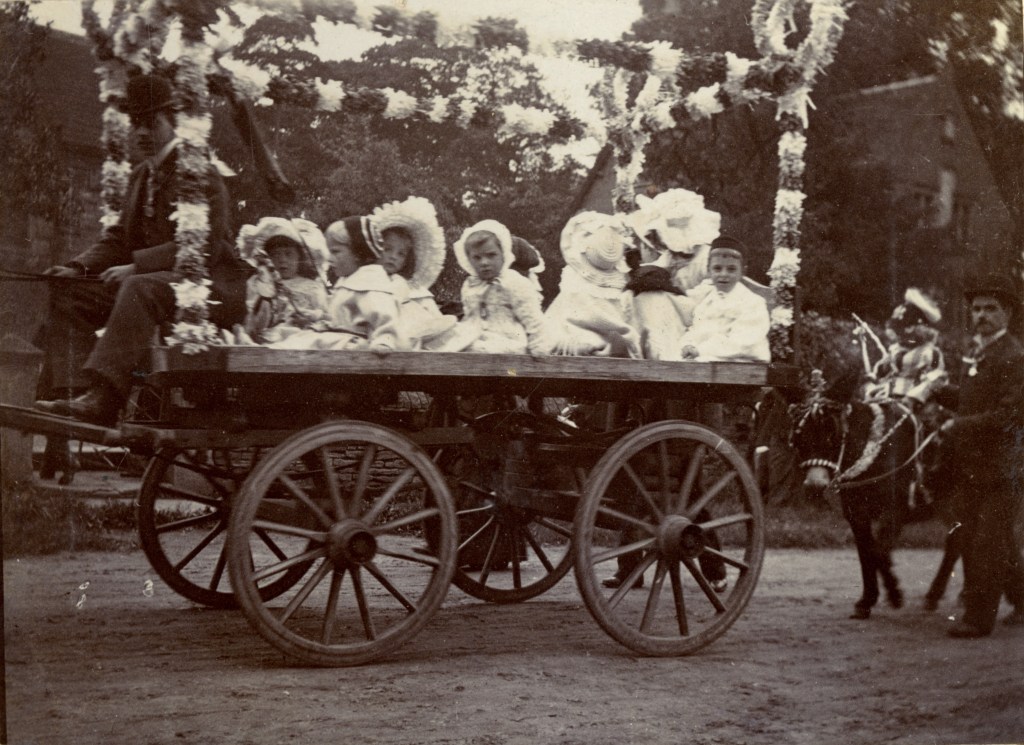 A historical photograph showing a horse-drawn cart decorated with flowers, carrying several children in traditional clothing during the Coronation Day celebrations in Cawthorne, 1902.