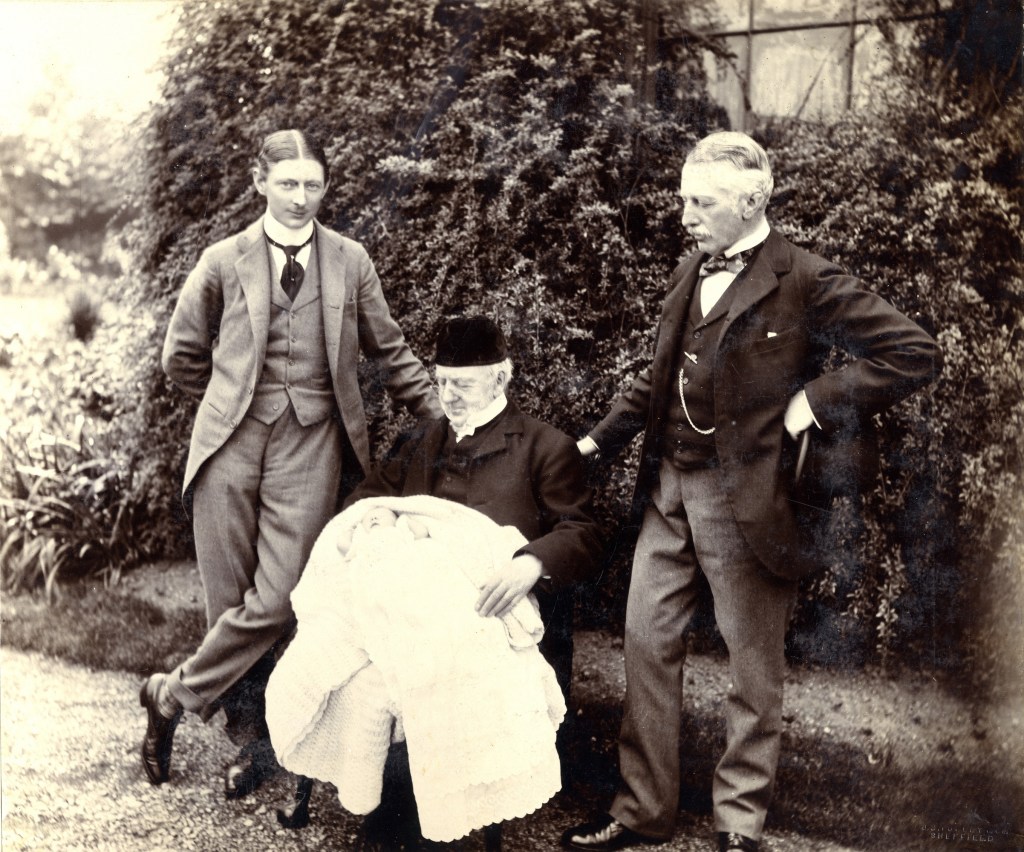 Three men in formal attire pose outdoors, one seated holding an infant wrapped in a white blanket, with greenery in the background.