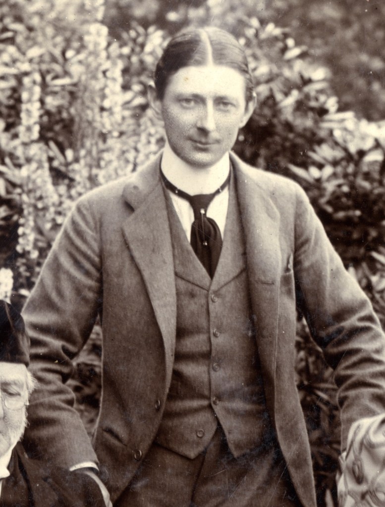 A young man in early 20th-century attire standing in a garden, with flowers in the background.