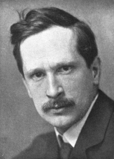 A black-and-white portrait of a man with a mustache, wearing a suit and tie, looking directly at the viewer.