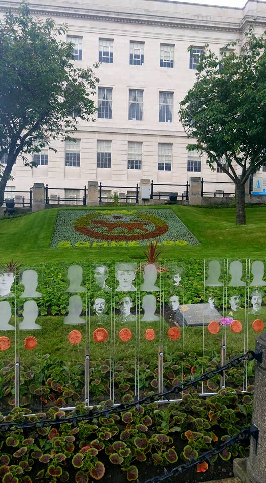 A beautifully arranged flower bed featuring a central emblem, with transparent panels displaying portraits of soldiers, located in front of a historic building.