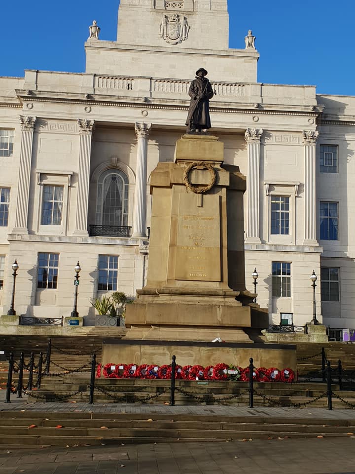 A war memorial featuring a statue of a soldier in uniform stands in front of a historic town hall building, adorned with wreaths at its base.