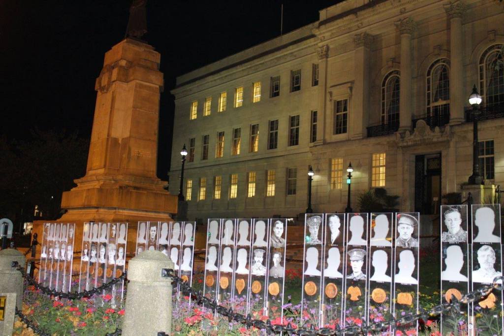 A night view of Barnsley's war memorial, featuring the statue atop the monument surrounded by illuminated portraits and floral arrangements in front of the town hall.