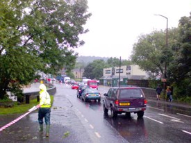 A flooded road scene with several cars and a police officer overseeing the situation during a rainstorm.