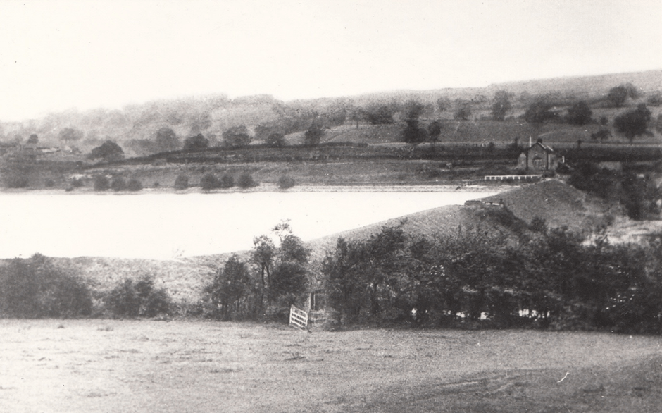 A historical black and white photograph depicting a scenic countryside view with a lake, hills in the background, and a distant mill structure, surrounded by trees and open fields.