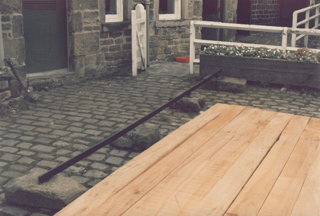 A cobblestone pathway featuring wooden planks and a metal bar, leading to a stone building with white railings and flower boxes.