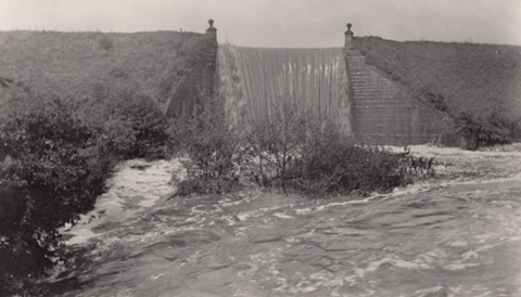 A historical black and white photograph depicting a dam with water flowing over it, surrounded by floodwaters and vegetation.