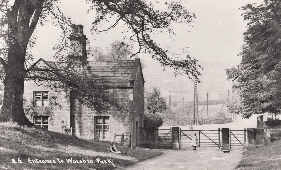 A historical photograph of the entrance to Worsbrough Park, featuring a stone building with a sloped roof, large trees nearby, and an open gate leading to the path.