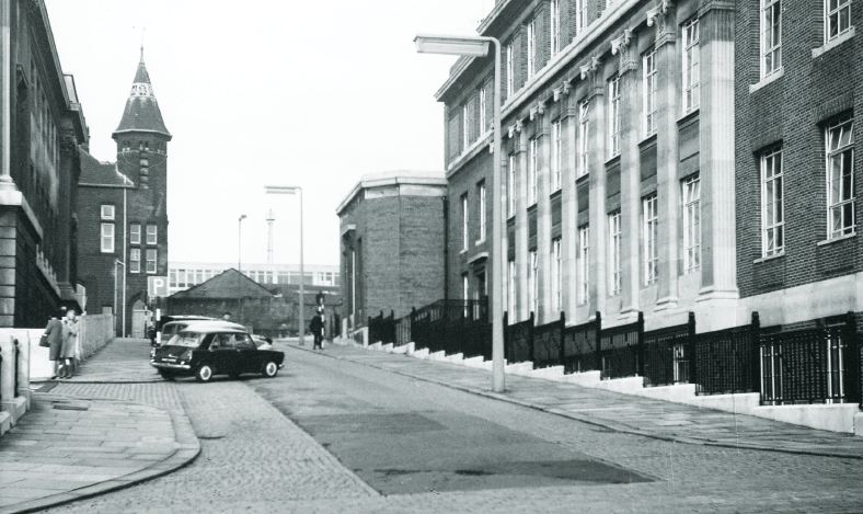 A black and white photograph depicting a street scene with a steep incline, featuring a parked car and pedestrians. Notable buildings line the street, showcasing architectural details of the early 20th century.