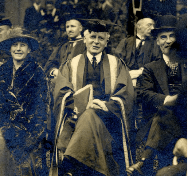 A historical black-and-white photograph of a group seated together, featuring a man in academic regalia in the center, surrounded by two women and other men in formal attire, likely at a university or formal event.