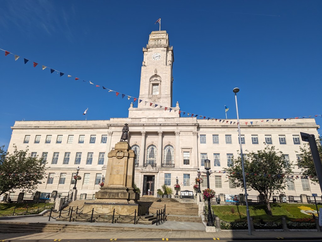 A view of Barnsley Town Hall with its clock tower, featuring a war memorial statue in the foreground. The scene is decorated with colorful bunting against a clear blue sky.