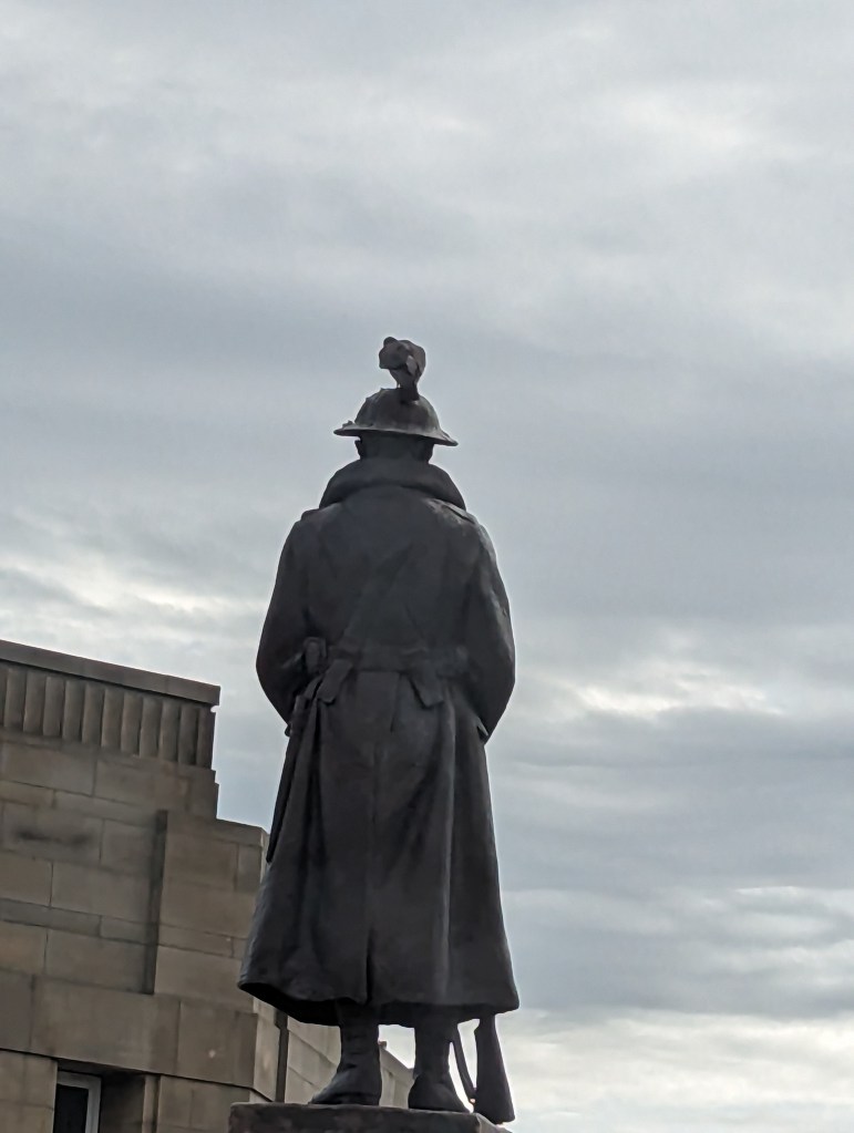 A bronze statue of a soldier from the back, wearing a military coat and helmet, with a pigeon perched on top, under a cloudy sky.