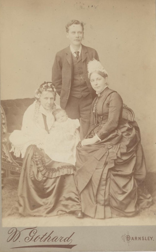 A historical black and white photograph featuring a young man in formal attire standing with three women, one of whom is holding a baby. The group is posed against a backdrop with ornate furniture, emphasizing Victorian style.
