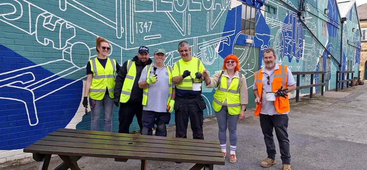 A group of six volunteers wearing high-visibility vests poses together outside against a mural backdrop, showcasing their enthusiasm for community work.