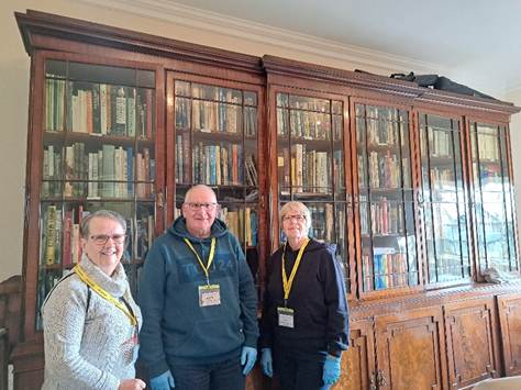 Three volunteers standing in front of a wooden bookshelf filled with books in a heritage site.