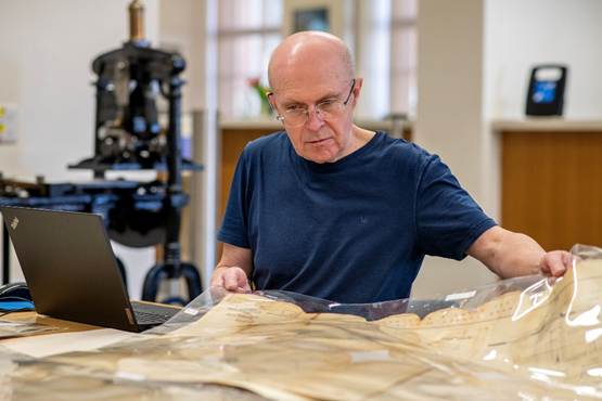 A volunteer examining historical documents at a desk with a laptop and archival materials in a museum setting.