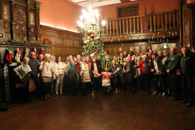 A large group of volunteers gathered indoors, joyfully posing together in front of a beautifully decorated Christmas tree, with festive decorations and smiling faces.