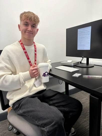 A young man with light brown hair is smiling while sitting at a desk holding a coffee cup. He is wearing a white sweater and black jeans, with a lanyard around his neck. A computer monitor is visible behind him, displaying a document.