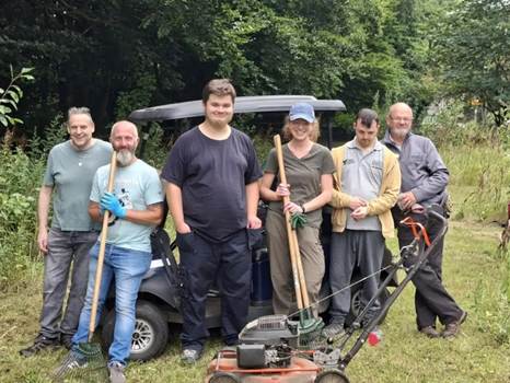 A group of six volunteers standing together in a grassy area, holding gardening tools such as rakes and spades, with a lawnmower in front of them and a utility cart in the background.