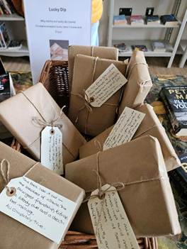 A basket filled with neatly wrapped brown paper parcels tied with string, each labeled with handwritten tags, in front of a sign for a 'Lucky Dip' at a second-hand bookshop.