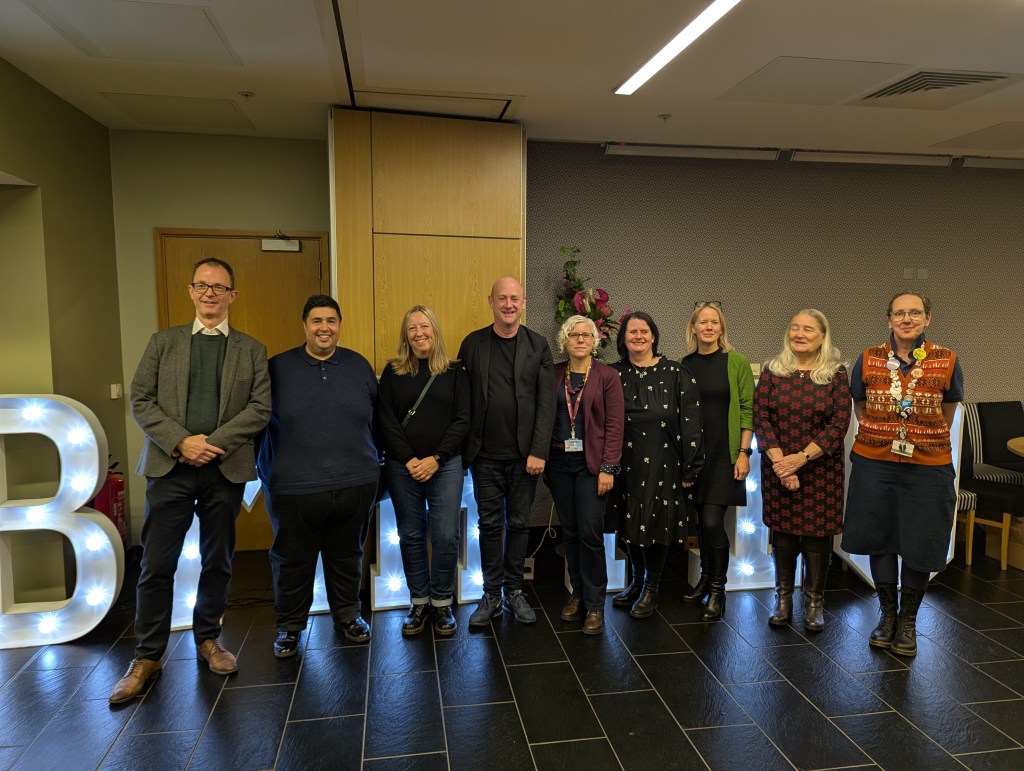 A group of ten individuals standing together in front of illuminated letters that spell 'BRIGHT'. The setting appears to be a modern indoor environment.