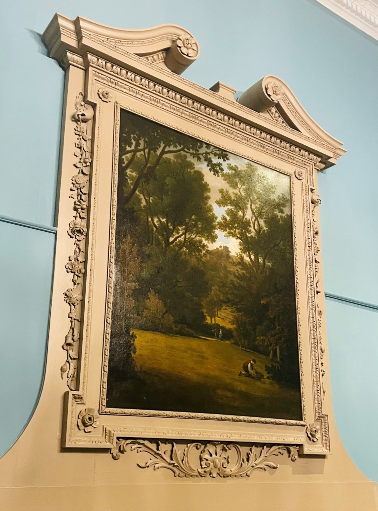 A framed painting of a pastoral scene featuring greenery and figures, displayed on a decorative wall.