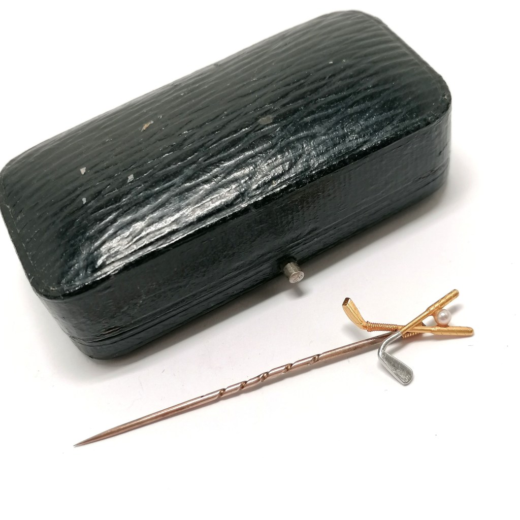 A vintage black case with a textured surface beside a gold and silver golf club motif tie pin, featuring a small pearl.