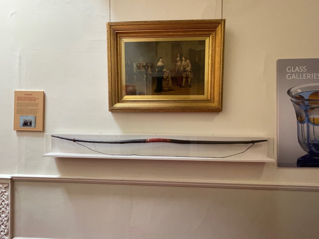 A longbow displayed underneath a framed painting in a gallery. The longbow is encased in glass on a shelf, with a descriptive plaque to the left and a poster on the right.
